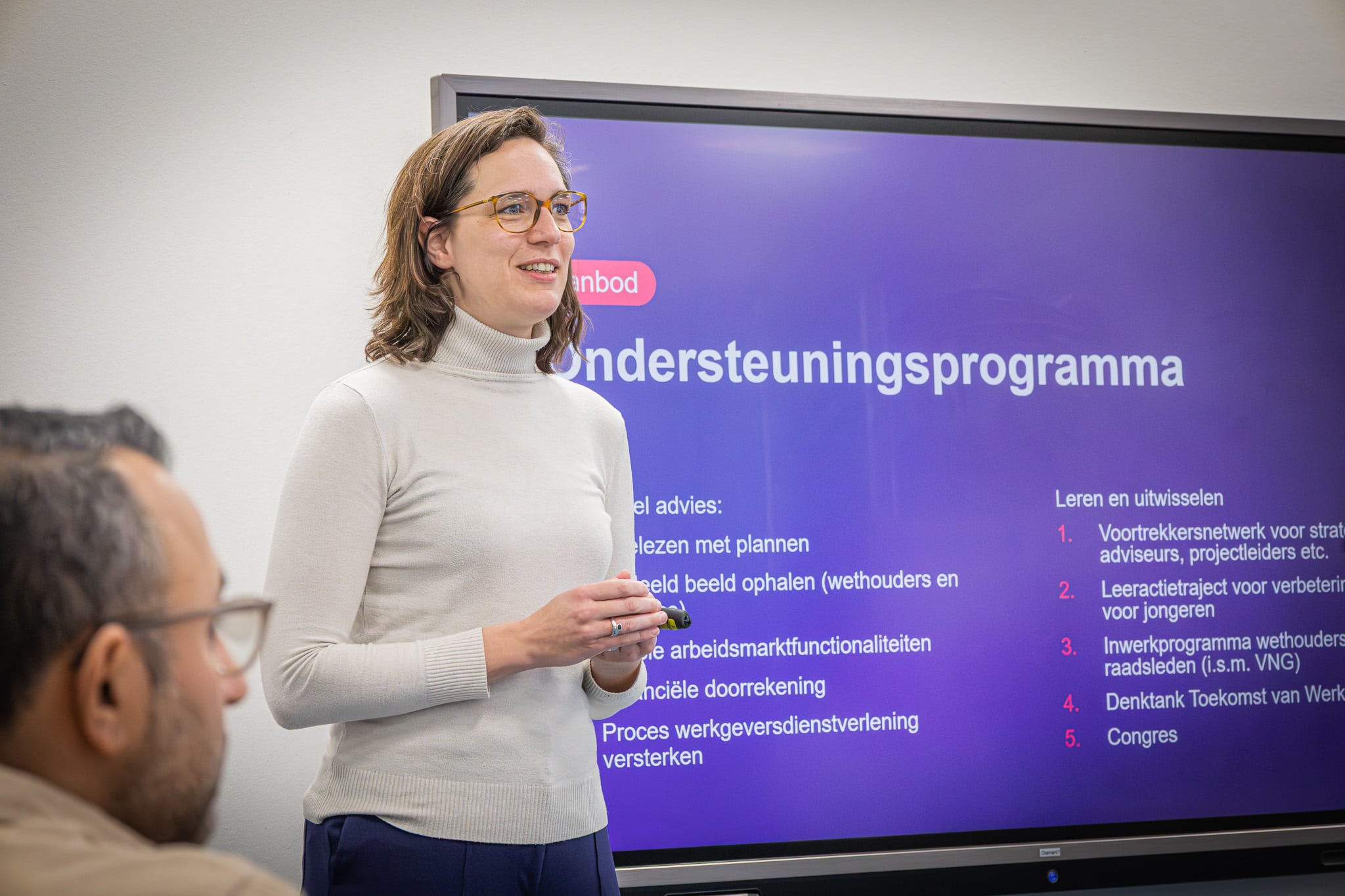 Marjolein van der Sterren geeft een presentatie tijdens een regionale bijeenkomst over de toekomstbestendige sociale infrastructuur. Op een groot scherm achter haar staat een overzicht van het ondersteuningsprogramma, terwijl deelnemers aandachtig luisteren.